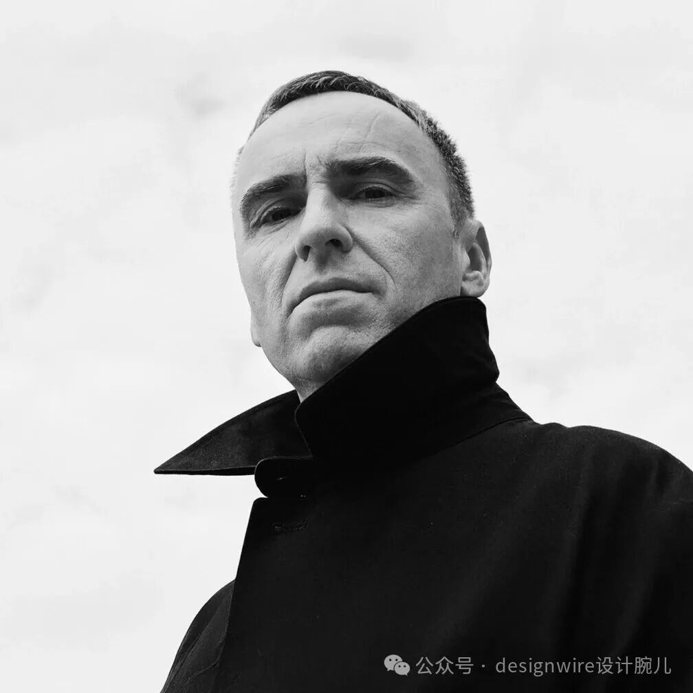 一张黑白低角度仰拍人像照片，画面主体为一名中年男性，短发，面部有明显皱纹，神情严肃，目光望向斜上方。他身穿一件高领黑色外套，衣领宽大且挺括，与浅色背景形成强烈对比。背景为均匀的灰白色天空或墙面，无其他杂物，突出人物轮廓。右下角有水印文字。