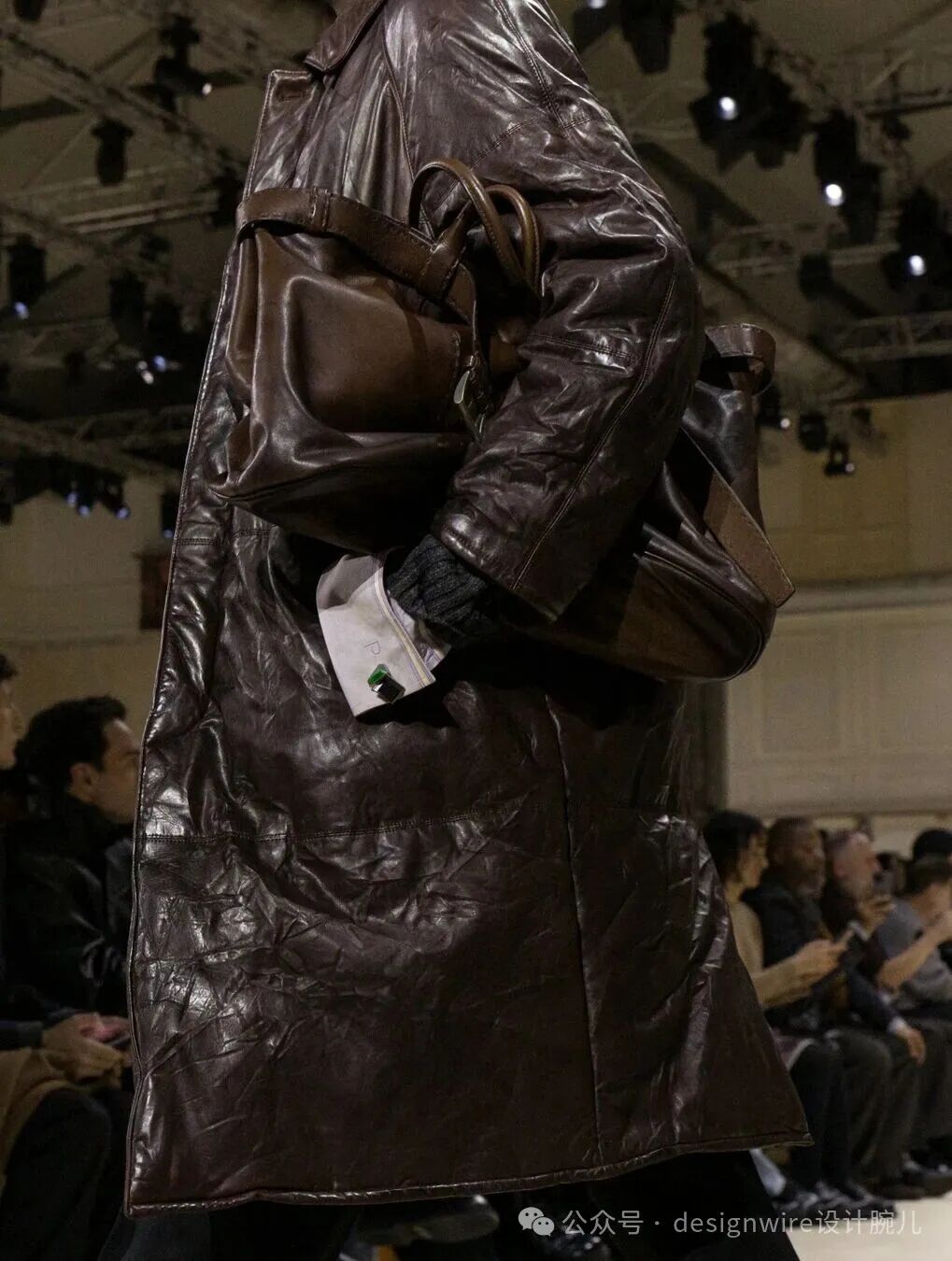 一位模特身穿深棕色褶皱皮革长款外套，外套材质具有明显光泽与自然褶皱纹理；右手提着同色系大号皮质手提包，包带宽厚，包身饱满；左手插在口袋中，露出白色袖口及黑色针织手套，袖口处有绿色与黑色相间的袖扣装饰；背景为室内秀场环境，可见顶部灯光设备及观众席上坐着的观众；右下角有水印文字“公众号 · designwire设计腕儿”