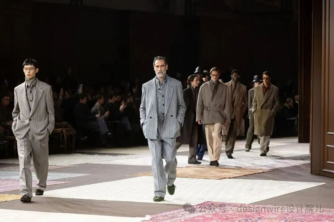 一场男装时装秀现场，多位男性模特在T台上依次走秀。前景左侧一位年轻亚洲男性模特身穿浅灰色竖条纹西装，内搭高领衫，双手插兜，戴眼镜；中央一位中年男性模特留灰白胡须，穿同款竖条纹双排扣西装套装，步伐稳健；其后方数位模特穿着不同色调的大地色系大衣与长裤，风格复古沉稳，部分佩戴帽子或墨镜。T台地面铺有几何拼接地毯，背景为昏暗观众席，可见多位观众就坐观看。画面右下角有水印文字“公众号 · designwire设计腕儿”。