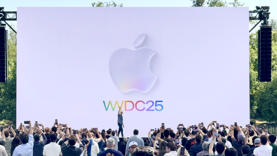 户外WWDC25活动现场,大屏幕显示立体浮雕苹果标志与WWDC25字样,演讲者举手示意,观众举手机拍摄