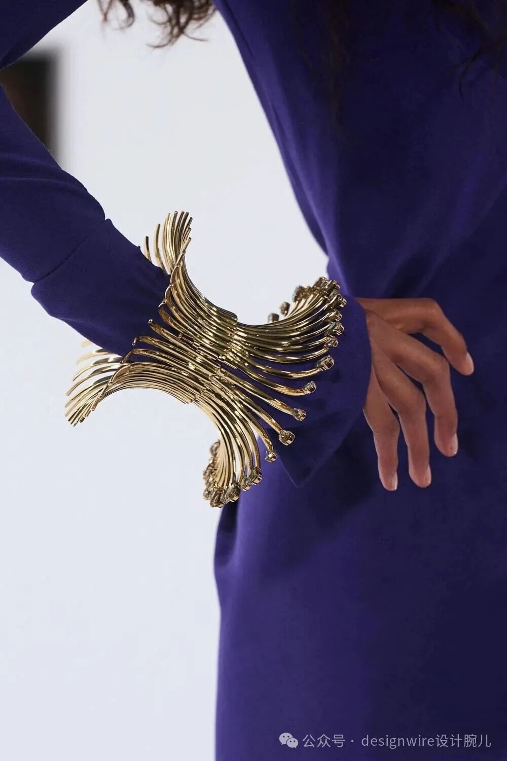 一位穿着深紫色长袖连衣裙的女性侧身站立，手臂自然下垂，手腕佩戴一件造型独特的金色宽版手镯。该手镯由多条细长弯曲的金属条组成，呈放射状或羽翼状展开，末端点缀有小颗透明宝石，整体设计富有动感与雕塑感。背景为纯白色，画面右下角有水印文字“公众号 · designwire设计腕儿”。
