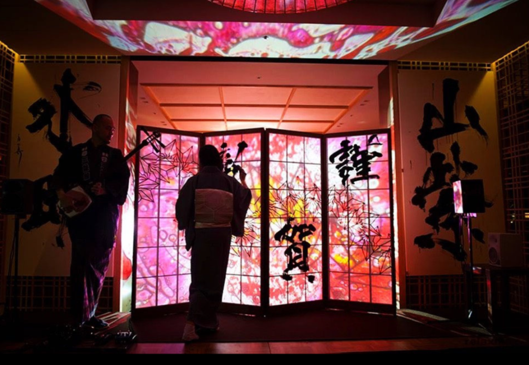 室内舞台场景，一名穿和服的背影人物站在可折叠的日式屏风前，屏风透出粉红与橙黄交织的抽象光影图案，屏风上写有黑色书法汉字。左侧一名戴眼镜、穿深色传统服饰的男子手持类似三味线的弦乐器正在演奏。舞台两侧墙面有大幅黑色书法字，左侧为“外”，右侧为“山”及下方竖排字；屏风上可见“謹賀”等字样。天花板投影着流动的粉红与白色抽象图案，整体光线偏暗，以暖色调为主，营造出日式传统与现代光影结合的表演氛围。