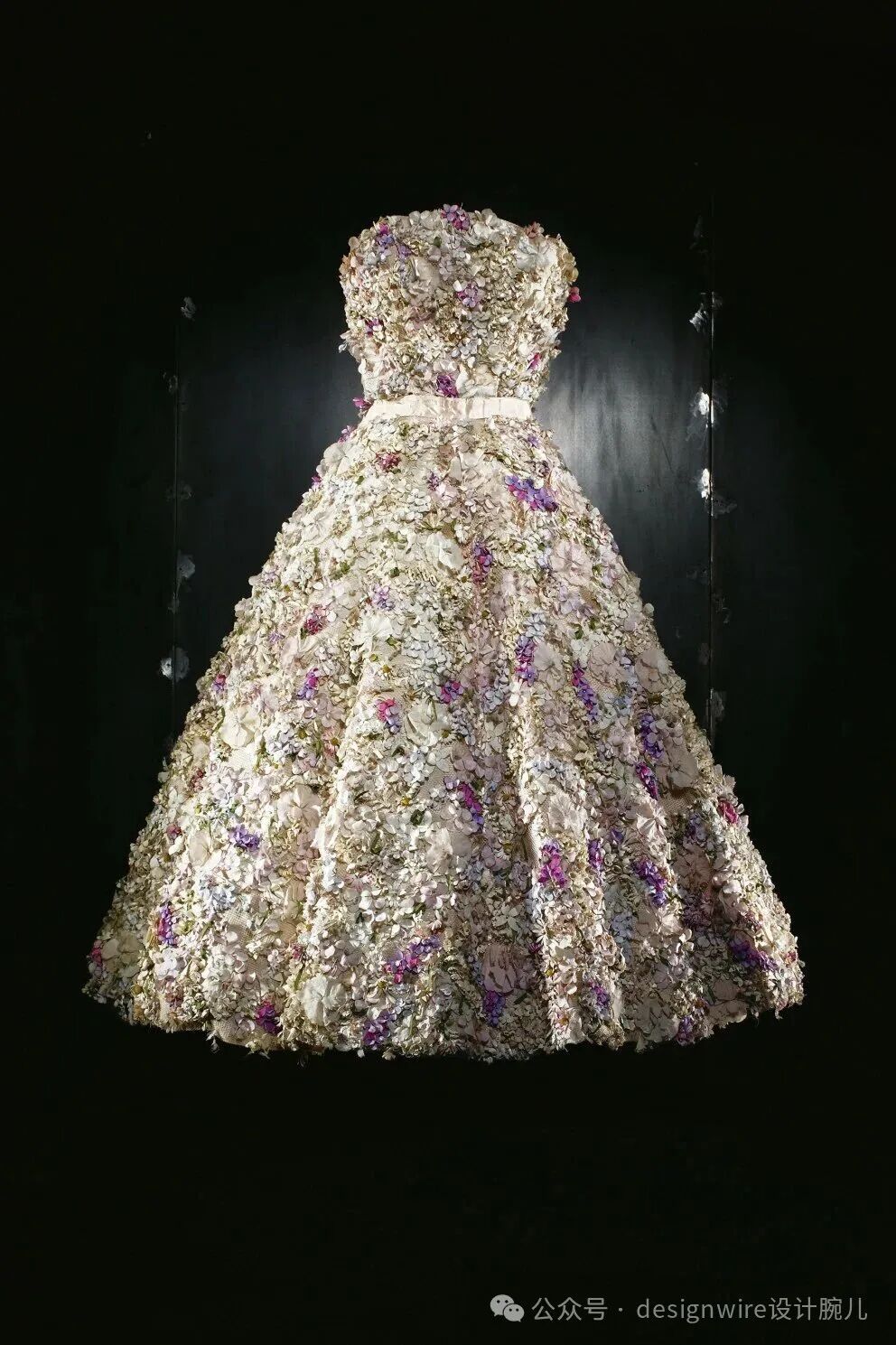 一件悬挂在黑色背景前的无肩带礼服，整体由大量立体花朵装饰而成，主要为白色、米色和紫色的花瓣，层次丰富，呈A字裙摆造型；腰部系有一条浅色宽腰带；礼服两侧可见细长的悬挂装置，背景有局部灯光照射形成光斑；右下角有水印文字“公众号 · designwire设计腕儿”