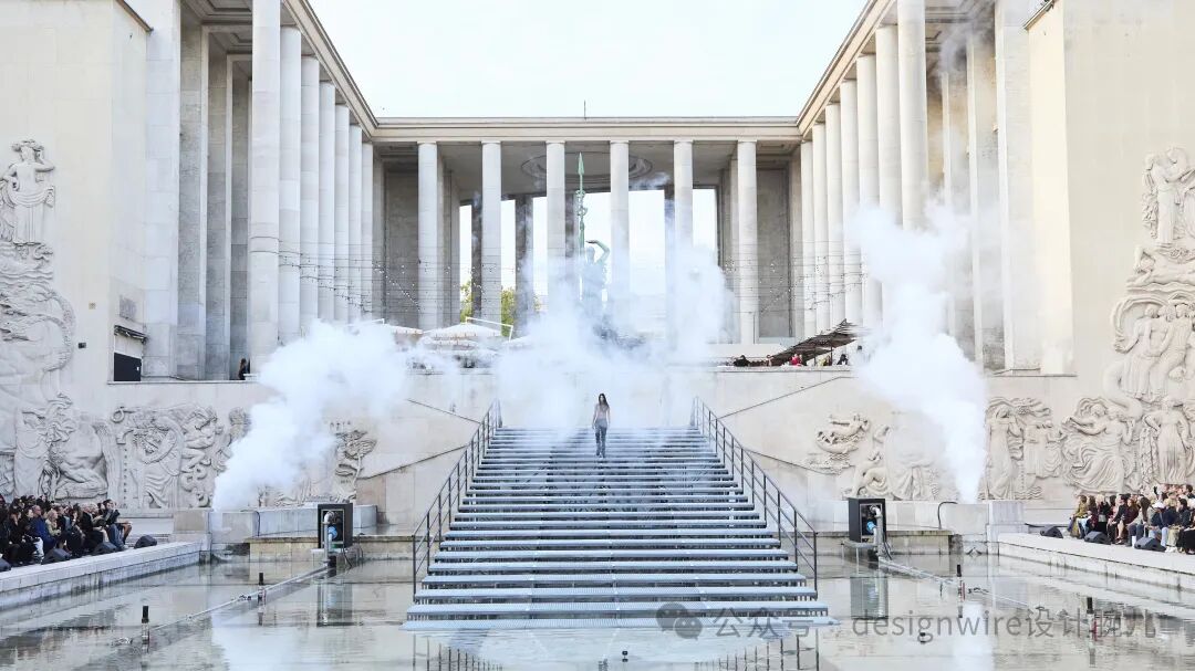 一座宏伟的古典风格建筑前，宽阔的石阶中央有一位穿浅色上衣和深色裤子的女性正沿台阶向上行走；台阶两侧及底部有大量白色烟雾喷出，弥漫在空中；台阶两侧设有金属扶手，下方是浅水池，水面倒映着台阶与烟雾；建筑立面布满高大圆柱与大型浮雕装饰；左右两侧台阶边缘坐有观众，部分人在拍照；画面右下角有水印文字“公众号：designwire设计网”；整体色调偏冷白，光线明亮均匀。