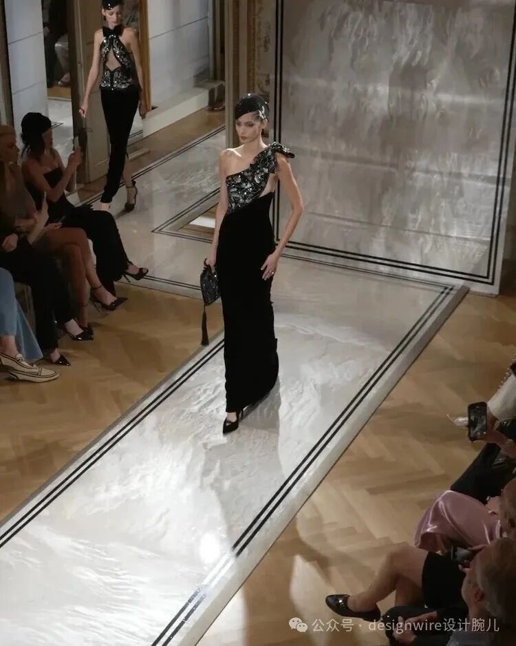 一位女模特在室内时装秀T台上走秀，身穿黑色单肩长裙，肩部饰有黑底银色亮片装饰的荷叶边，搭配黑色高跟鞋与手拿包，头戴黑色发饰；T台为白色带双黑边的长条形，地面为木质拼花地板；两侧观众席坐有数人，部分正在拍照；背景可见另一位穿黑银相间上衣与黑裤的模特正从后方走来；右下角有水印文字“公众号·designwire设计腕儿”
