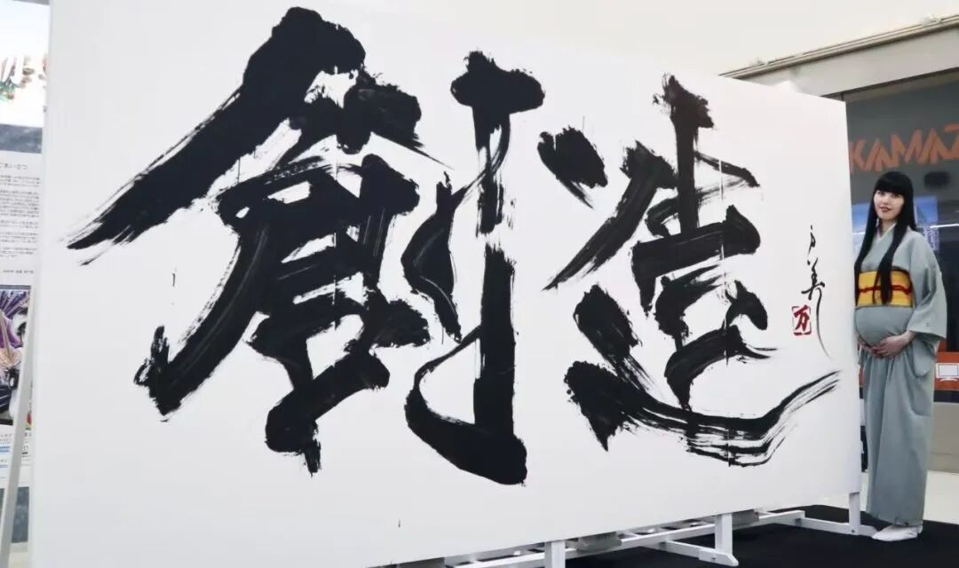 一名身穿浅灰色和服、腰系黄红相间宽腰带的孕妇站在一幅大型白色书法作品旁，双手轻抚腹部，面带微笑。书法作品以浓墨重彩的毛笔挥洒出两个大字“創造”，笔触奔放有力，墨色浓淡相宜，带有飞白与墨滴效果；右侧有红色印章及竖排小字落款。背景可见室内展览环境，左侧有展板。