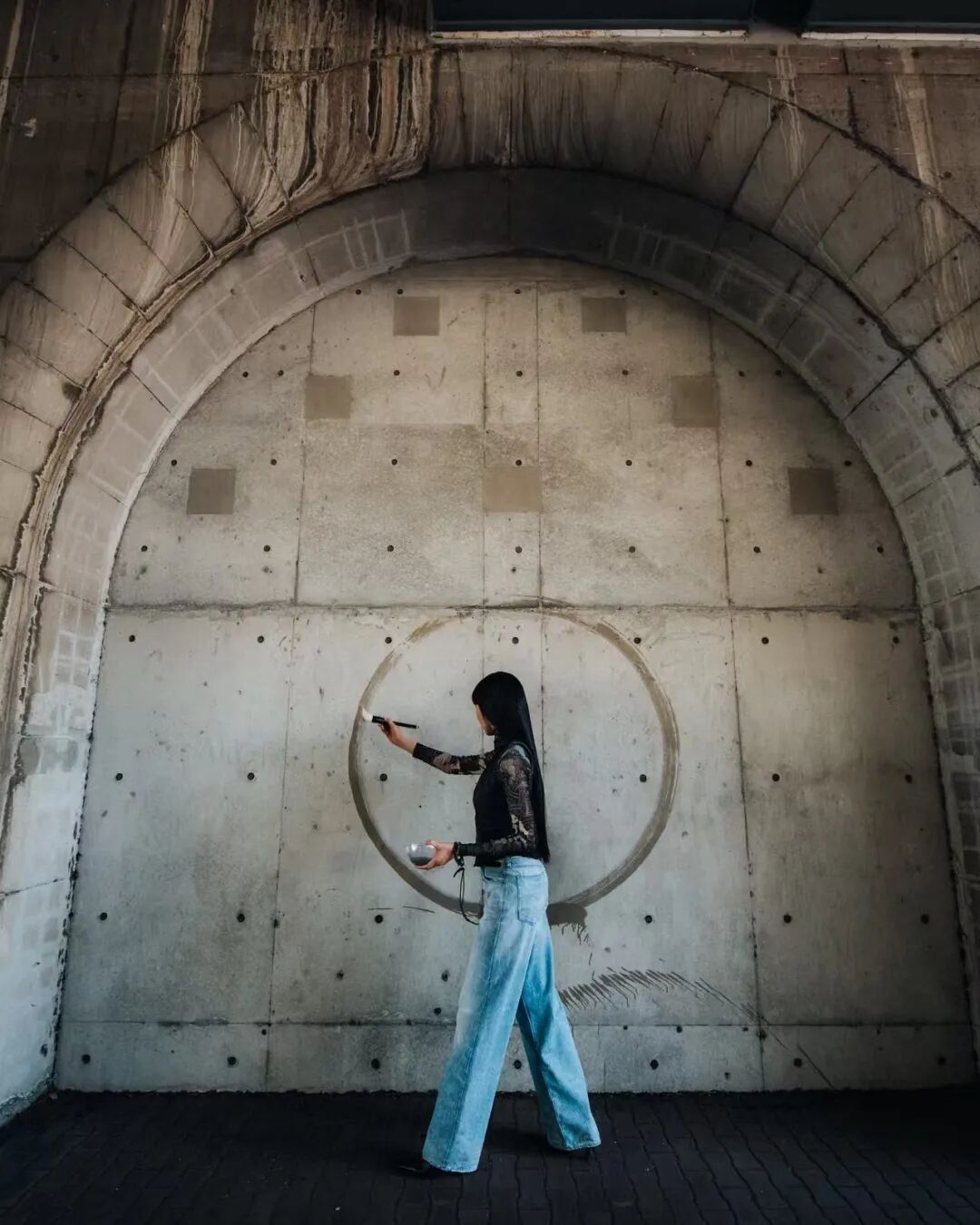 一名黑发女性站在混凝土拱形墙前，身穿黑色蕾丝长袖上衣与浅蓝色阔腿牛仔裤，右手持画笔正于墙面绘制一个大型圆形墨迹，左手托着盛有颜料的容器；背景为带有方形修补痕迹和均匀分布螺栓孔的灰色混凝土墙面，上方可见拱形结构边缘及垂直滴落的污渍痕迹，地面为深色砖块铺就，整体光线偏暗，呈现街头艺术创作场景。