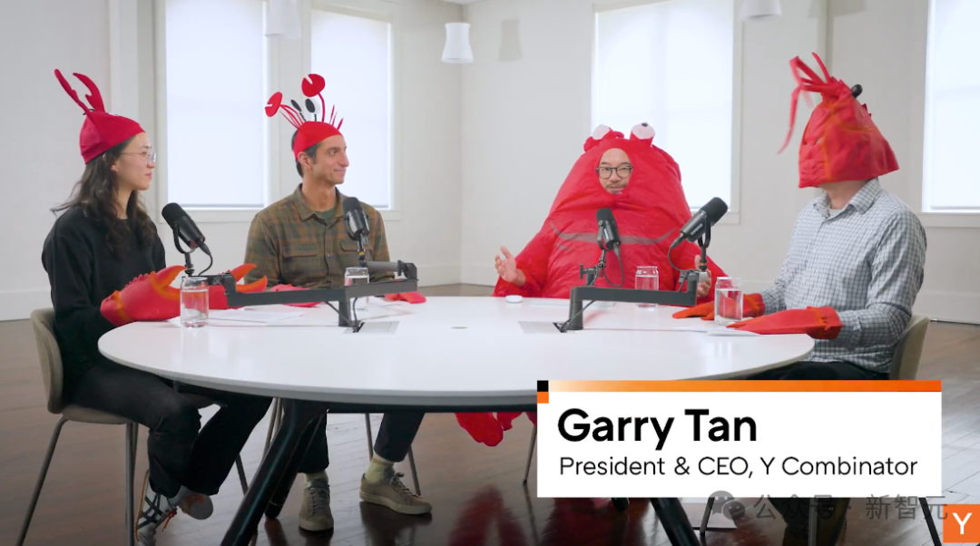 四个人围坐在一张白色圆桌旁，均戴着红色龙虾造型的头饰和手套。左侧女性戴眼镜，穿黑色上衣；中间男性穿格子衬衫；右侧一人穿着完整的红色龙虾连体服，戴眼镜；最右侧男性穿格子衬衫，头戴龙虾帽。桌上摆放多个麦克风、水杯和纸张。背景为明亮室内，有窗户和吊灯。