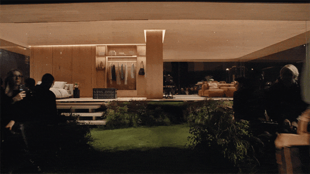 夜晚户外时装秀场景，以现代风格室内家居环境为背景，设有床、衣柜等家具，前方是绿色草坪T台。多张图片为动图抽帧，显示一名戴帽子、穿宽松外套与长裤的模特从左至右沿草坪走秀，手持包袋；两侧坐有观众，部分观众举着手机拍摄。整体色调偏暖黄，灯光聚焦于T台和背景建筑结构，营造沉浸式秀场氛围。