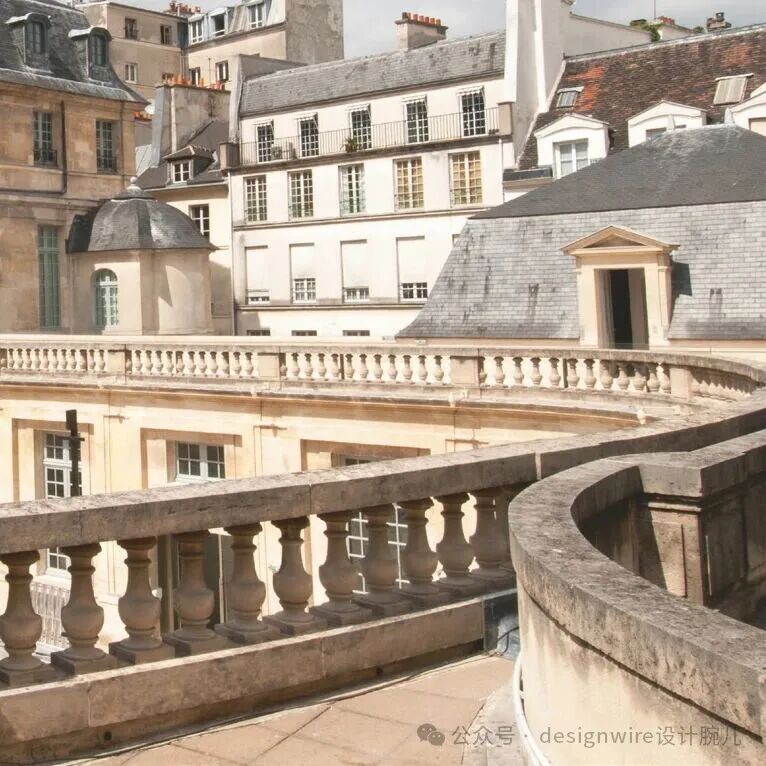 一张展现欧式古典建筑群的俯瞰视角照片，前景为石质弧形栏杆，由一排带有柱头装饰的立柱组成；中景是另一层弧形阳台栏杆，材质与前景一致；背景是多栋米黄色或浅色外墙的法式风格建筑，屋顶多为深灰色瓦片，可见多个老虎窗、烟囱及带铁艺护栏的窗户；画面右下角有水印文字‘公众号 designwire设计腕儿’，整体色调偏暖黄，光线柔和，呈现宁静典雅的城市街景氛围。