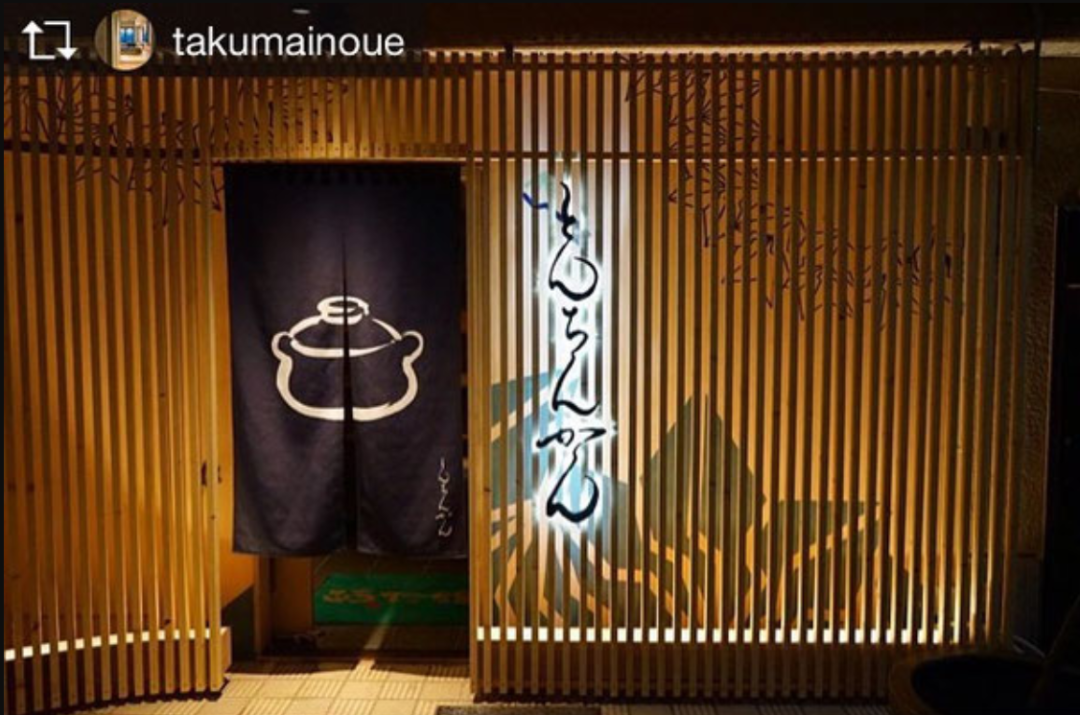 夜晚拍摄的日本传统餐馆入口，木质竖条格栅门面，左侧悬挂深蓝色布帘，上有白色陶锅图案及小字；右侧格栅上以发光效果呈现日文书法“もんじゃ”（Monya），背景隐约可见绿色植物剪影；整体暖光照明，地面为浅色地砖。