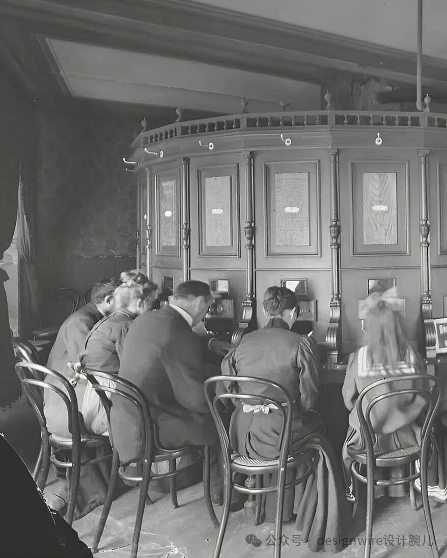黑白历史照片，展示一间室内场景：数人背对镜头坐在弯曲的木质操作台前，操作台呈半圆形，设有多个带玻璃窗的隔间，内部可见设备或文件；人们穿着20世纪早期服饰，包括西装、长裙与围裙；座椅为金属框架的弯椅（如Bistro椅）；背景有花纹墙纸、吊灯及装饰性栏杆；右下角有水印文字‘公众号 · designwire设计腕儿’。