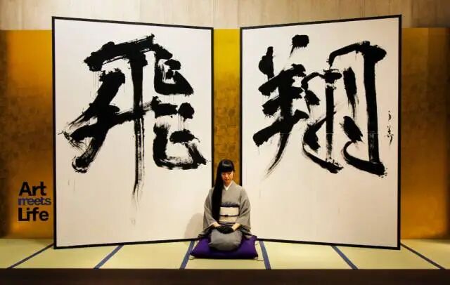 一名身穿灰色和服、腰系白色宽腰带的女性盘腿坐在紫色坐垫上，位于两幅大型立轴书法作品之间。左侧立轴书写黑色大字“飛”，右侧立轴书写黑色大字“翔”，均为浓墨重彩的毛笔狂草风格，笔触奔放有力。背景为暖黄色木质墙面，地面铺有榻榻米，带有深蓝色条纹。