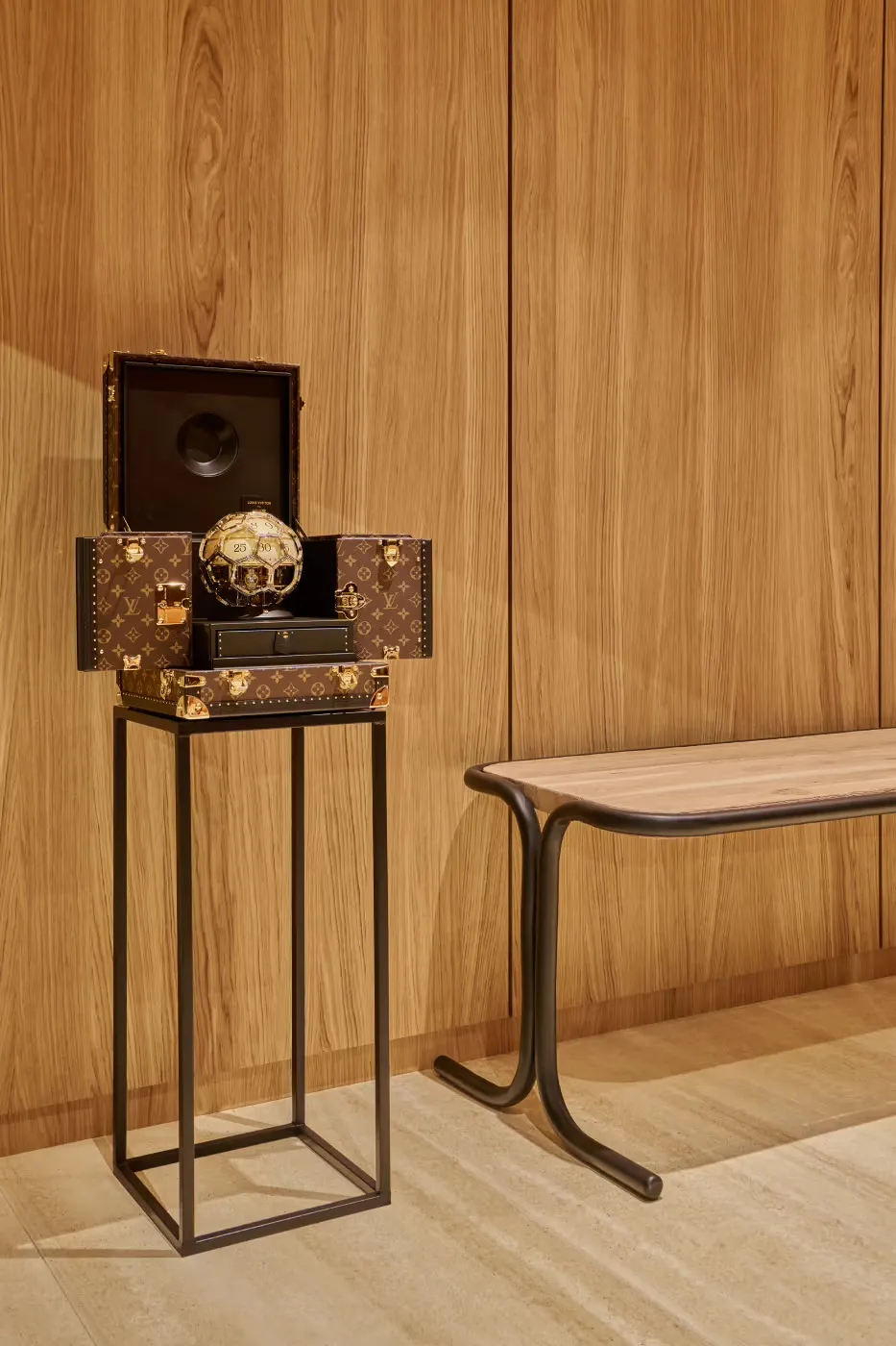 木质纹理墙面背景前，一个黑色金属方框支架上叠放着三个路易威登（Louis Vuitton）经典老花图案的硬质箱包：最上方为打开的大型手提箱，内有黑色衬里及圆形凹陷；中间是横向放置的中号箱包，其上摆放着一个金色足球造型装饰品；下方为小型箱包，顶部带锁扣。右侧有一张木面金属腿长凳，地面为浅色大理石纹瓷砖，整体呈现高端奢侈品陈列场景。