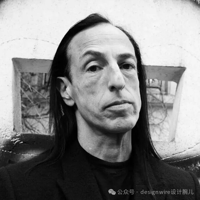 一张黑白肖像照，一位长发男性，头发乌黑且垂至肩部，面部有明显皱纹，鼻梁高挺，眼神望向侧前方。他身穿深色高领上衣和外套，背景为粗糙墙面，墙上有一个不规则的窗洞或装饰性开口，透过开口可见模糊的室外景物。照片右下角有水印文字。