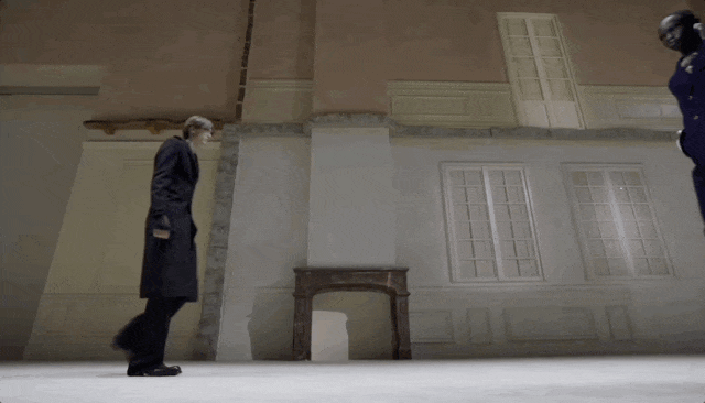 该系列图片为时装秀场走秀动图抽帧。第一、二张图显示两位模特在模拟建筑背景的T台上并行走秀，左侧为金发男性模特，身穿深色长款大衣；右侧为黑人男性模特，身穿深蓝色长款大衣并佩戴黑色面罩，两人步伐同步，背景有仿旧墙面、窗格及一张古典木桌。第三、四张图为同一男模（戴帽、穿黑色西装套装）沿T台正向走来，左手提黑色手袋，两侧坐满观众，场景为室内秀场，地面铺白，墙壁有做旧裂痕设计，整体风格冷峻、现代且具戏剧性。