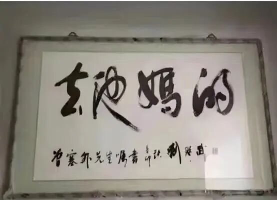 一幅装裱在白色边框内的书法作品，悬挂在浅色墙壁上，画心为白色宣纸，上有黑色毛笔行草书大字‘去他妈的’，下方有较小的落款文字‘曾寒外先生嘱书’及印章，整体风格粗犷有力，带有现代网络语境下的反叛或调侃意味。