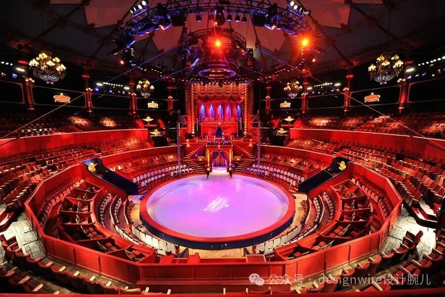 一个大型马戏团室内剧场的俯瞰视角，中央是一个圆形表演舞台，舞台地面呈淡紫色并带有白色图案，边缘为深蓝色；舞台后方是装饰华丽的红色帷幕舞台背景，上方悬挂着多组专业舞台灯光设备，包括聚光灯和彩色灯效；观众席呈环形阶梯式分布，座椅为红色木质结构，部分区域设有黑色滑梯状通道；顶部有吊灯和金属桁架结构，整体灯光以红色和紫色为主，营造出热烈而梦幻的氛围；右下角有水印文字“公众号：designwire设计腕儿”