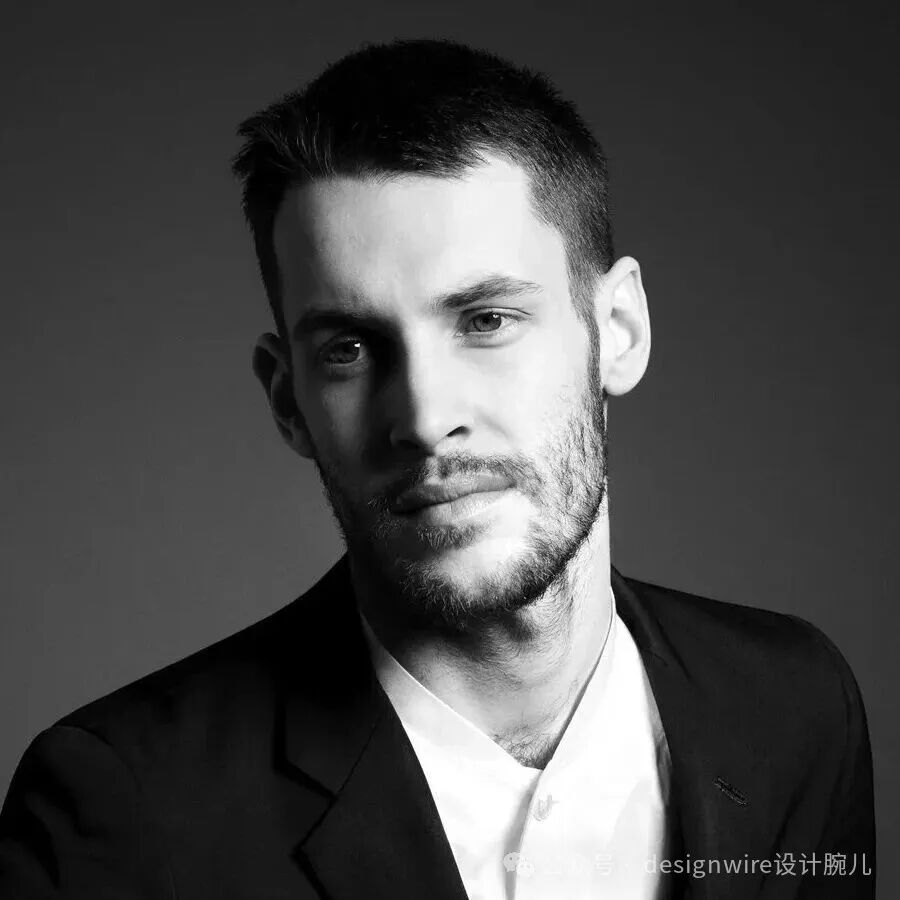 一张黑白风格的男性半身肖像照，男子留着短发和修剪整齐的胡茬，身穿深色西装外套搭配白色衬衫，领口敞开，目光正视镜头，表情沉静。背景为纯灰色渐变，光线从左侧打来，在面部形成明暗对比，突出轮廓。右下角有水印文字“designwire设计腕儿”及部分模糊图标。