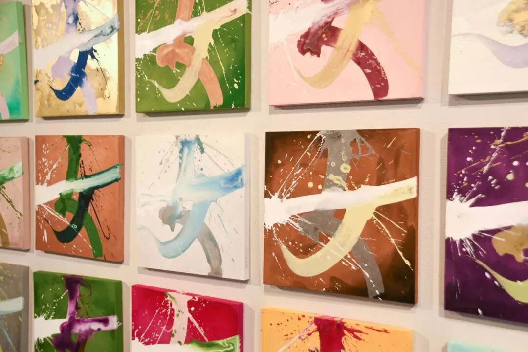 墙上陈列多幅方形抽象画作，每幅画以不同背景色为底（如绿色、粉色、橙色、紫色、红色、黄色等），画面主体为大笔触的泼墨或飞溅式书法笔画，常见白色、蓝色、金色、酒红色、灰绿色等色彩，部分画作有飞溅的颜料点与动态线条，整体呈现现代抽象艺术风格，画布呈网格状排列于浅色墙面上。