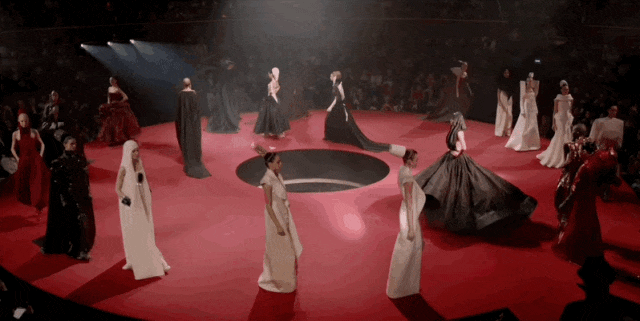 多帧动图抽帧展示一场高端时装秀场景：圆形红色T台中央设有一个黑色椭圆凹陷区域，多位模特身着黑白红三色长款礼服（含拖地长裙、斗篷式设计）在台上走秀或摆姿，部分模特手持道具；舞台上方多束白色聚光灯从不同角度照射，形成明亮光柱；观众席环绕T台，呈暗色背景；整体氛围华丽、戏剧化，具有强烈的视觉冲击力。