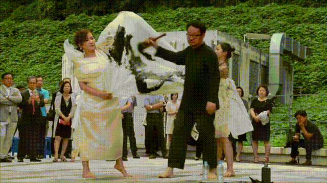 户外广场上进行的一场融合传统水墨与现代舞蹈的表演。画面中央一名穿黑色长衫、戴眼镜的中年男性赤脚，手持毛笔在白色大幅宣纸上挥毫泼墨，宣纸随动作翻飞；他两侧各有一名穿白色长裙的女性舞者，同样赤脚，配合其动作舒展肢体，其中左侧女舞者身着纯白长裙，右侧女舞者裙摆有水墨点缀。背景为绿色植物墙和一栋浅色建筑，前方围有观众，部分人鼓掌，右侧有蹲姿摄影者。地面可见水瓶与墨瓶等道具。整体呈现动态、富有张力的艺术表演场景。