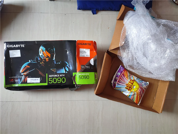 一张俯拍图片，展示了未拆封的技嘉RTX 5090显卡包装盒及旁边较小的橙色技嘉包装与打开的快递箱