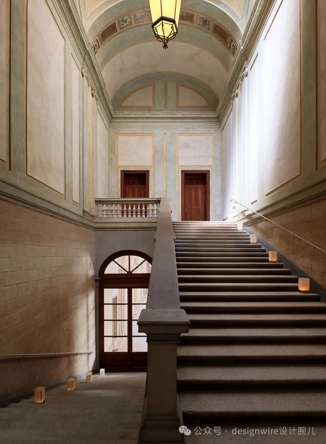 一座古典风格的室内楼梯间，中央为宽大的石质台阶，铺有深灰色地毯，两侧设有金属扶手；楼梯左侧是砖石墙面与一扇带拱形窗格的木门，右侧墙面为浅色护墙板，上方设有一处带栏杆的二层平台，平台后方有两扇对称的木门；顶部为拱形天花板，饰有几何与花卉浮雕图案，中央悬挂一盏黄铜框架的复古吊灯；楼梯台阶及地面散置多个方形纸灯笼，散发暖黄色光晕，营造出庄重而温馨的氛围。