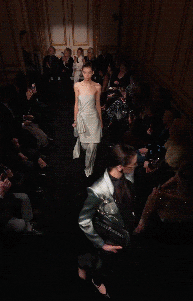 一组时装秀现场照片，展示多位模特在室内T台走秀。场景为古典风格的室内空间，墙壁有金色镶边装饰，灯光聚焦于T台。前景和中景有多位女性模特依次走过，主要穿着浅灰或米白色无肩带长裙，裙身有褶皱、系带或不对称设计，搭配同色系长裤或高跟鞋；其中一位模特手持银色手包。观众席坐满人群，多人举手机拍摄。画面因动态抓拍存在轻微模糊，整体色调偏暗，突出服装光泽与模特姿态。