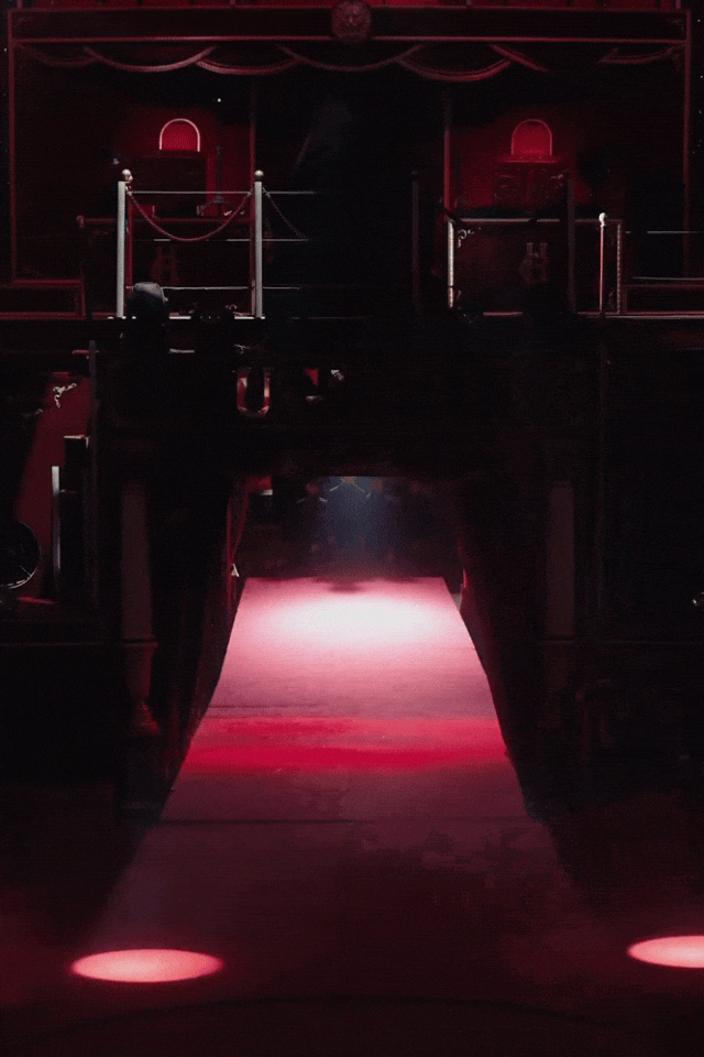 一场舞台场景的静态图像，整体色调为深红色与黑色，氛围神秘。画面中央是一条被聚光灯照亮的红色地毯通道，从前景延伸至中景的拱形门洞。通道两侧有装饰性立柱支撑的横梁结构，横梁上刻有文字。背景是一个带有红色帷幕、金色边框和波浪形顶饰的舞台布景，上方悬挂一个狮子头造型的装饰物。舞台左右对称设有带栏杆的平台，平台上放置有道具箱（印有字母H）、灯具及类似鼓或提篮的物品。地面可见两个圆形红色光斑，光线主要来自上方和前方，形成强烈的明暗对比。图像具有轻微噪点与动态模糊感，疑似动图抽帧。