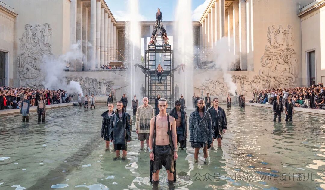 一场户外时装秀场景：多位模特站在大型水池中，水位及膝，中央一位赤裸上身、穿破洞短裤的男性模特正对镜头；其后方有数位穿着深色宽松外套或无袖服饰的模特；背景是古典风格建筑，两侧有高大立柱与浮雕墙面，中央设有一座黑色金属阶梯结构，顶部与中间平台各有人物站立或坐姿；多处喷泉向上喷水，水雾弥漫；两侧水池边缘聚集大量观众；画面右下角有水印文字“公众号 - designwire设计腕儿”