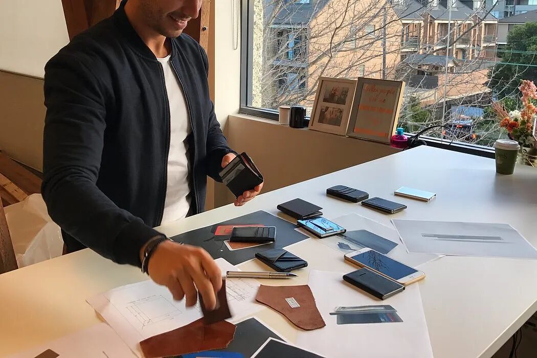 一名穿着黑色夹克和白色内搭的男子坐在桌前，手中拿着一个黑色钱包