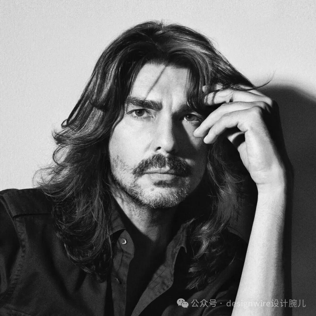 一张黑白人物肖像照，一位留着中长卷发、有胡须的男性，身穿深色衬衫，右手轻触额头，目光直视镜头，背景为纯浅色墙面，光线从左侧打来，在面部和头发上形成明暗对比，右下角有水印文字。