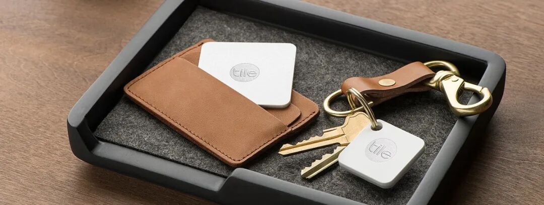 一个黑色托盘内放置着一个棕色皮质钱包、一把钥匙和两个Tile追踪器