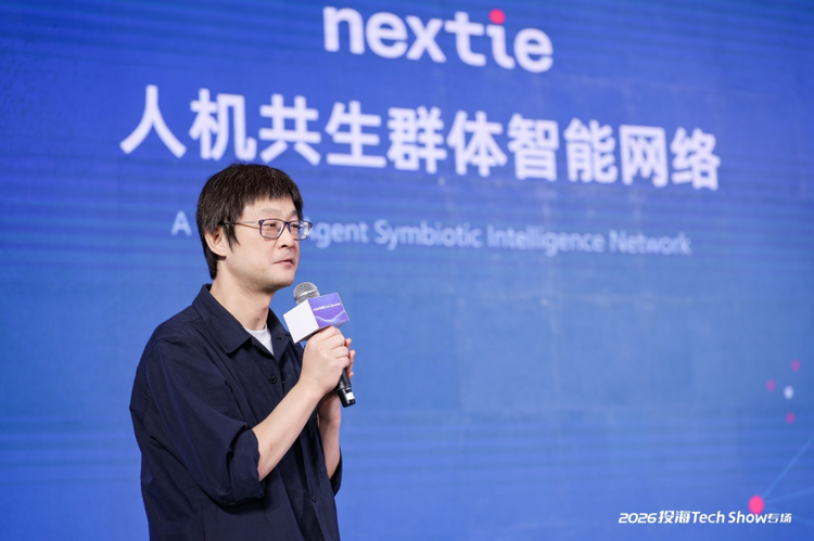 一位戴眼镜的中年男性站在蓝色背景板前演讲，背景显示“人机共生群体智能网络”