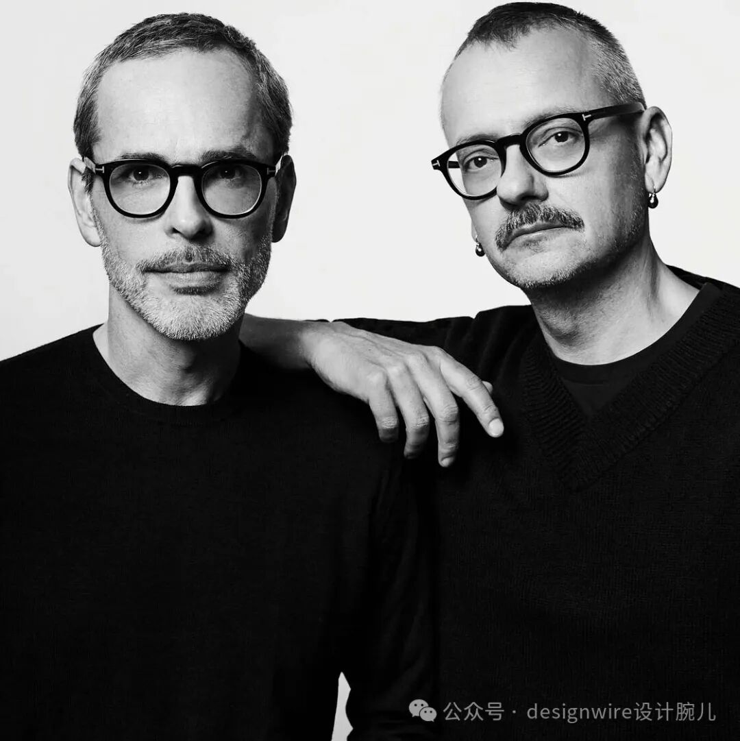 一张黑白风格的双人肖像照，画面中两位中年男性并肩站立，背景为纯白色。左侧男子短发灰白，留有山羊胡，佩戴黑框眼镜，身穿黑色圆领上衣；右侧男子发型较短、头顶微秃，有浓密上唇胡须，同样佩戴黑框眼镜，左耳戴有黑色耳环，身穿黑色V领毛衣，右手搭在左侧男子肩上，手指自然伸展。整体构图简洁，光线柔和均匀，突出人物面部轮廓与表情。右下角有水印文字。