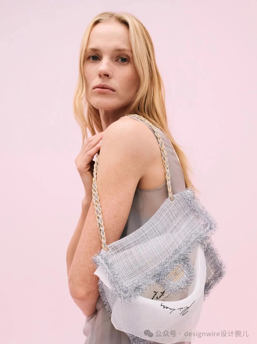 一位金发女性侧身站立，背景为纯粉色。她身穿浅灰色无袖上衣，肩上斜挎一个设计独特的手提包：包身主体为白色半透明材质，外覆一层蓝白细格纹织物，边缘有毛边处理；包带为金色链条样式。包的下部可见黑色手写体签名式文字，右下角有水印文字‘公众号 · designwire设计腕儿’。