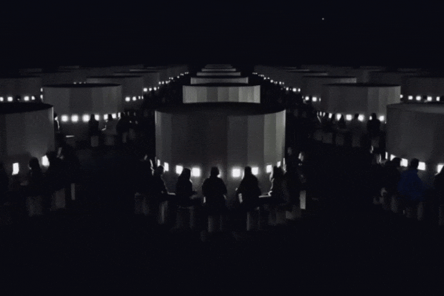 夜间户外场景，多排大型圆柱形白色结构体呈行列状排列延伸至远方，每个结构体侧面嵌有发光的方形灯带，形成明亮的光带效果。结构体周围及前方聚集着许多人的剪影，他们或坐或站，面向结构体，部分人坐在低矮的圆形凳子上。画面整体为黑白灰调，背景是纯黑夜空，右上角可见一颗微小的亮星。图像具有轻微动态模糊感，似为动图抽帧，人物与灯光位置略有变化，但整体布局稳定。