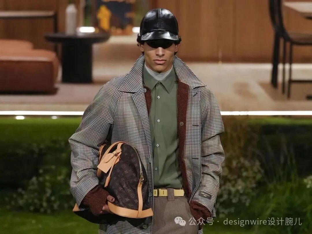 一名男性模特在T台走秀，头戴黑色亮面皮质棒球帽并佩戴黑色墨镜，身穿灰白格纹长款外套，内搭橄榄绿色衬衫与白色高领打底衫，外套内衬为酒红色针织材质；腰间系米色宽版皮带，下身搭配灰色长裤；左手持一个深棕色老花图案的LV背包，包带与饰件为浅棕色皮革；背景为室内布景，含木质家具、黑色圆桌、橙色沙发及绿植装饰；画面右下角有水印文字“公众号・designwire设计腕儿”