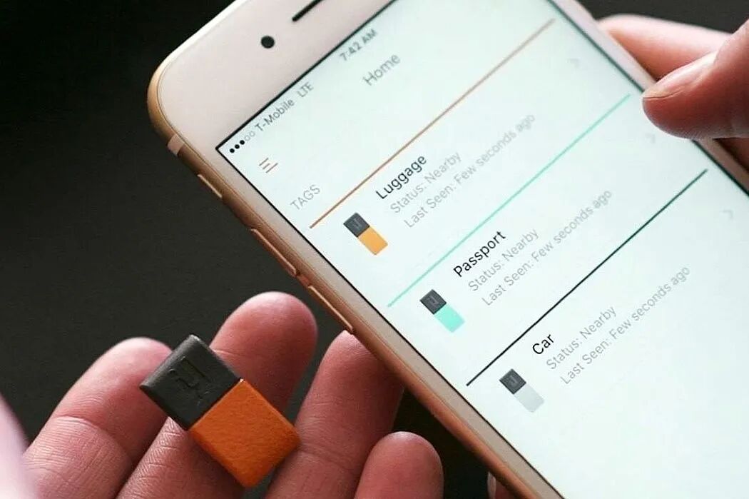 一只手拿着一个橙黑相间的迷你追踪器，另一只手握着一部显示追踪应用界面的智能手机