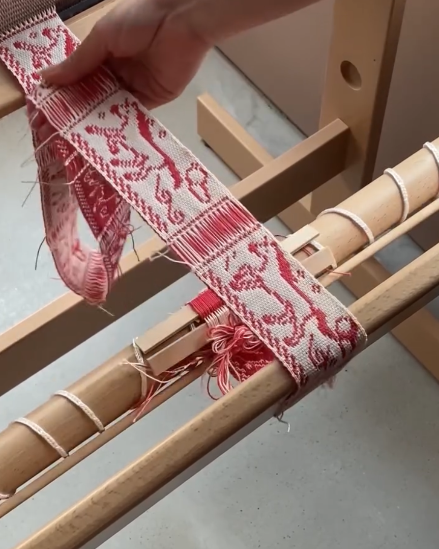 手工织布机特写：木制框架，红白相间编织带，图案为传统动物纹样