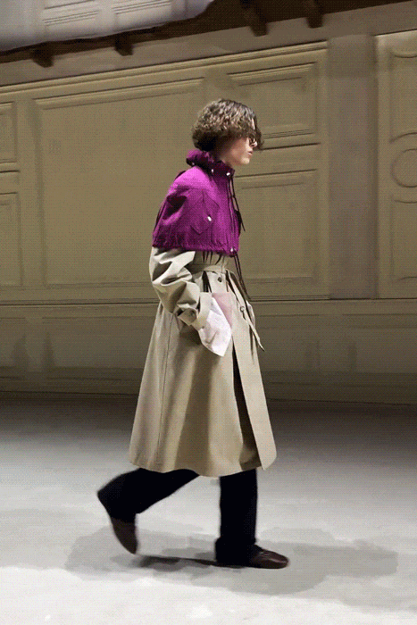 图片展示了一场男装时装秀的多个瞬间：第一张图中，一位卷发模特身着紫色高领针织衫搭配米色长款风衣、黑色长裤与深色皮鞋，正侧身行走；第二张图中，另一位模特穿着红色无袖针织上衣与黑色长裤，手插口袋行走；第三、四张图为同场秀的群像镜头，多位不同肤色与发型的年轻男性模特排成一列前行，服装色彩丰富，包括绿色连帽衫、粉色束腰外套、灰色西装等，部分模特佩戴帽子或手持白色手套，背景为浅色带有浮雕装饰的墙面。