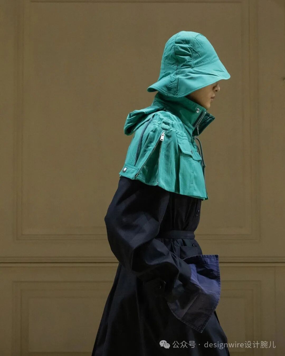 一位侧身行走的模特，身穿深蓝色长款风衣，外搭一件青绿色短款连帽夹克，夹克领口高耸并带有拉链和口袋设计；头上戴着同色系宽檐渔夫帽，帽檐下遮住部分面部；背景为浅米色带凹凸线条的墙面；画面右下角有水印文字。