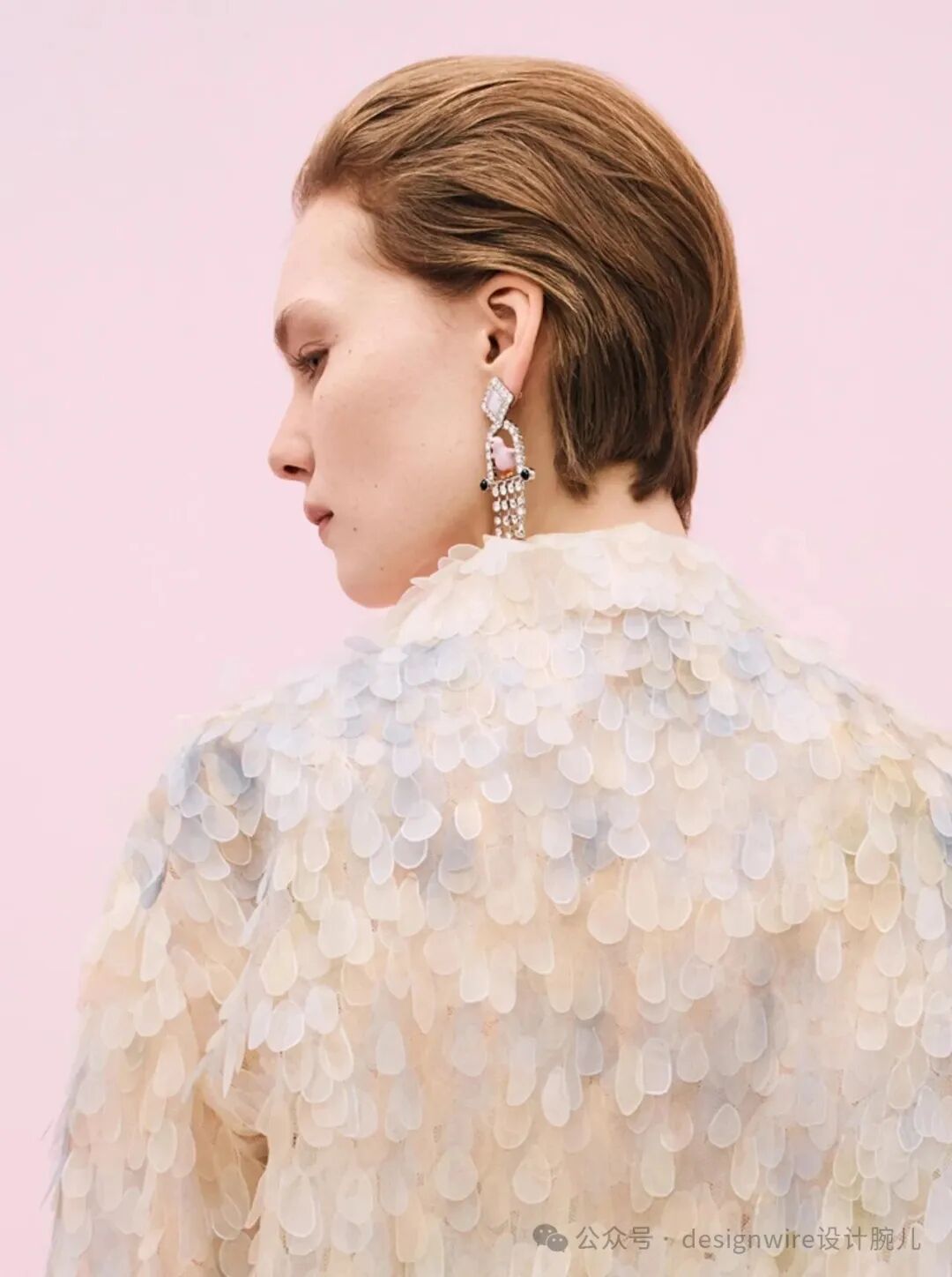 一位女性侧身背对镜头，留着棕色短发，发型整齐贴合头部。她佩戴着一只华丽的长款耳环，耳环上部为几何切割钻石造型，中部嵌有粉色调宝石，下部垂挂多排闪亮水晶珠串。她身穿一件由半透明、水滴形片状材料（类似贝壳或树脂）层层叠叠覆盖的高领礼服，材质轻盈通透，呈现淡雅的米白、浅蓝与奶油色渐变。背景为纯粉色，整体画面柔和梦幻。右下角有水印文字“公众号 · designwire设计腕儿”。
