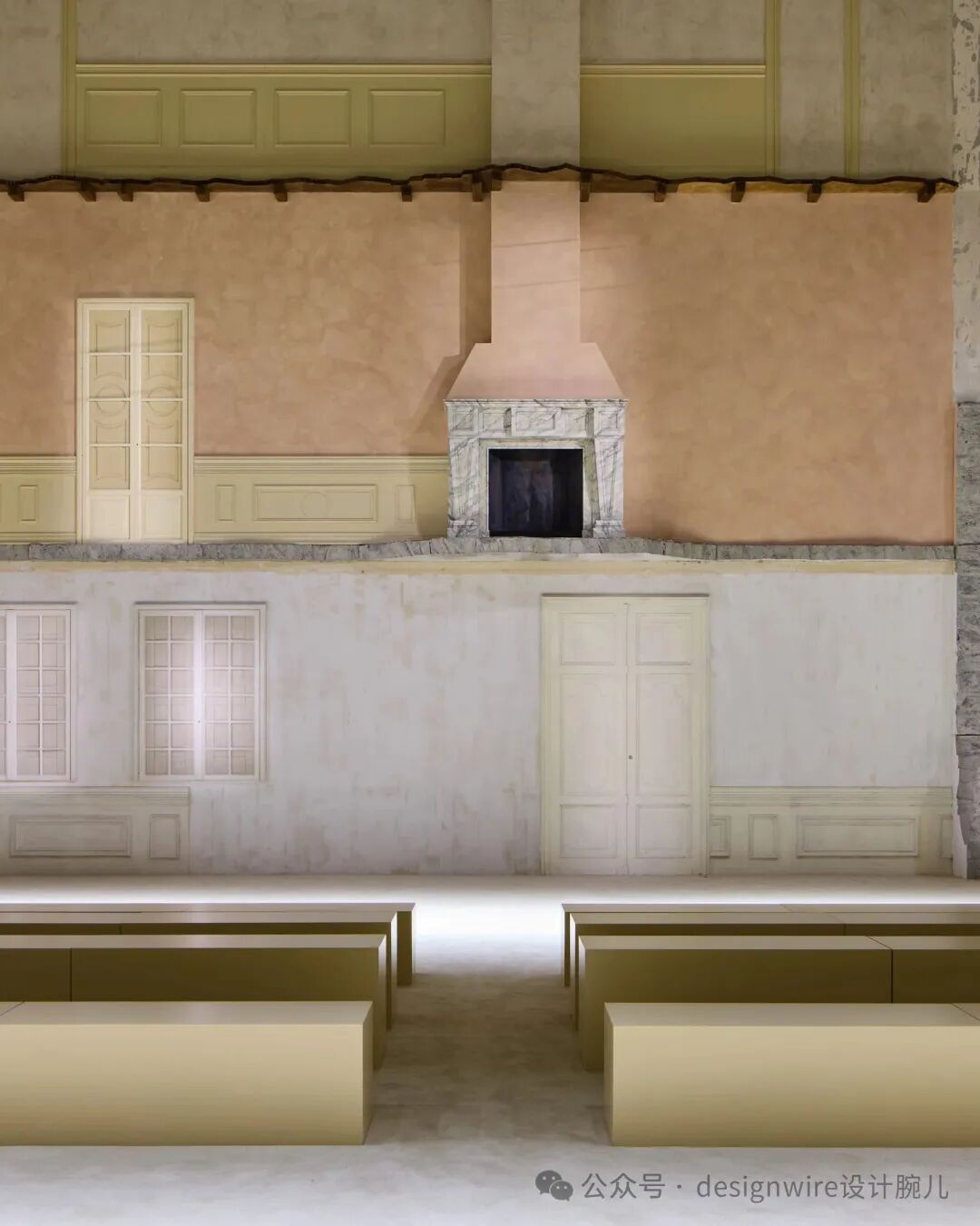 一张展示室内空间设计的图片，画面呈现上下分层结构：上半部分为粉橙色墙面，中央设有一个白色大理石边框、粉色烟囱罩的壁炉，左侧有一扇米白色双开门；下半部分为浅灰白色墙面，设有两扇带格子窗的窗户和一扇米白色双开门，墙下有装饰性护墙板；前景是几排浅米色长条形座椅或展台，地面为浅色材质；右下角有水印文字‘公众号 · designwire设计腕儿’。