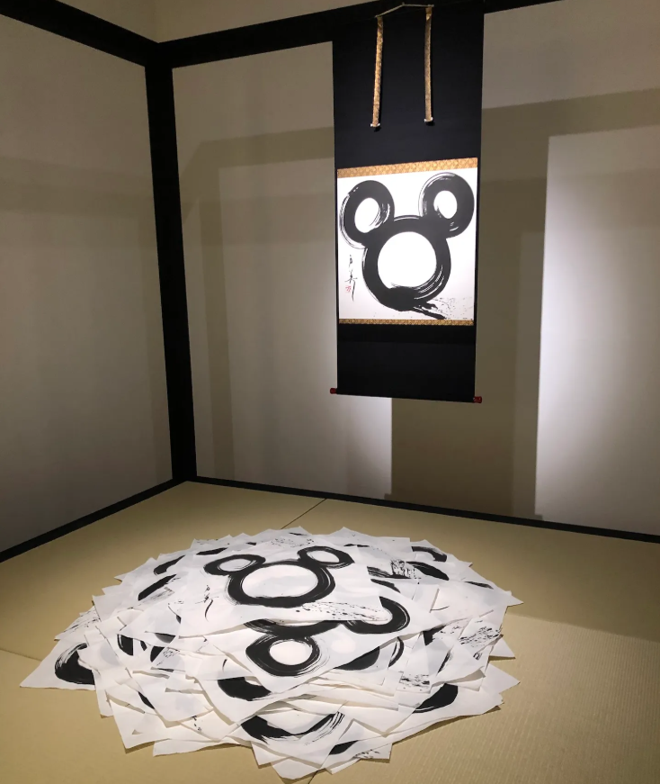 室内场景，日式风格房间角落，铺有榻榻米，墙角为黑边白墙。墙上悬挂一幅黑色卷轴，卷轴中央为白色底，绘有以水墨技法创作的米老鼠头像轮廓（两个圆耳加一个大圆脸），画作上下有金色装饰边框，右侧有红色印章及落款书法；卷轴由两根浅棕色绳索悬吊。地面中央堆叠着大量散乱的白色纸张，每张纸上均印有或手绘相似的黑色水墨米老鼠头像图案，部分纸张边缘有墨迹晕染与笔触痕迹，整体呈现艺术展览或创作过程中的作品展示状态。