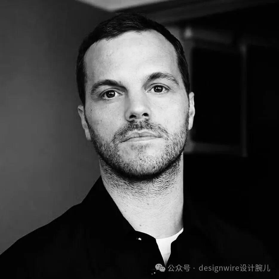 一张黑白风格的男性面部特写照片，人物为短发、有胡茬，身穿深色衬衫内搭白色衣领，目光直视镜头，背景为模糊室内环境，右下角有水印文字‘公众号 · designwire设计腕儿’