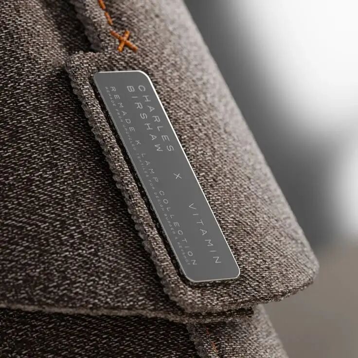 灰色纹理织物上的长条形金属标签，印有CHARLES BIRSHAW x VITAMIN COLLECTION等品牌信息