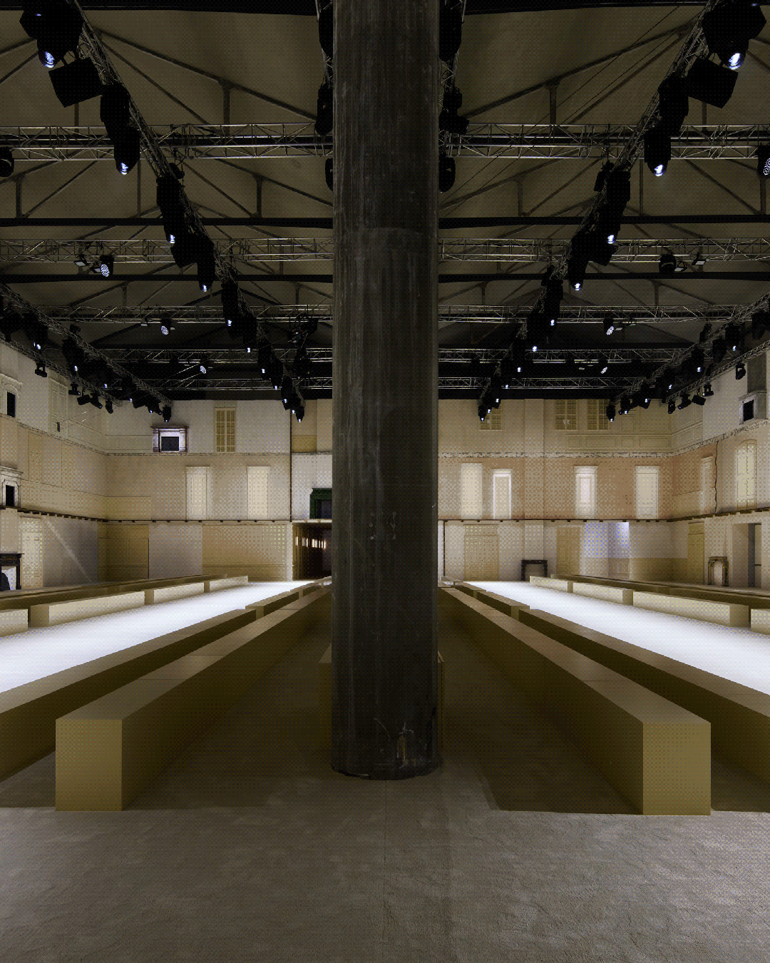 一个大型室内秀场或展览空间，中央有粗壮的深灰色混凝土柱子，两侧对称排列着浅米色长条形阶梯式观众席或展示台，地面为浅灰色。天花板是高挑的工业风格钢架结构，悬挂着大量黑色专业舞台射灯。背景墙面为复古风格，呈现米黄色与浅褐色拼接，带有古典壁板、窗户和门洞造型；其中一张图清晰显示了壁炉与烟囱结构。整体光线明亮均匀，色调偏暖黄，营造出庄重而现代的氛围。