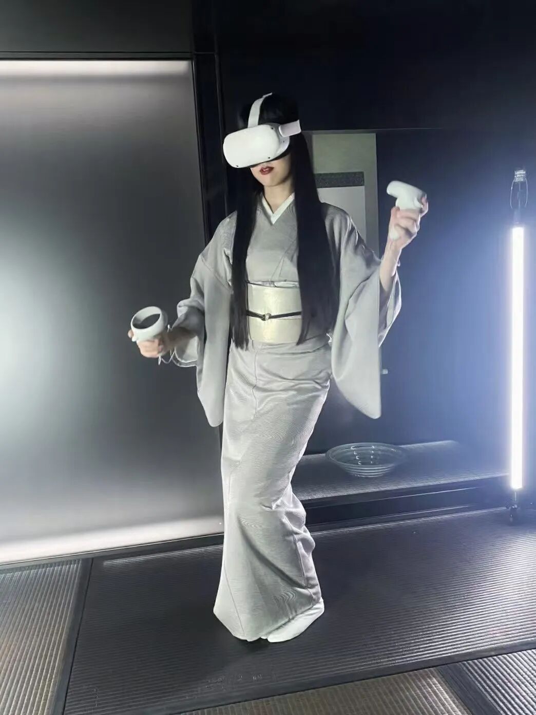 一位长发女性身着浅灰色传统和服，腰间系有银色宽腰带，佩戴白色VR头显设备，双手各持一个白色VR控制器，呈站立姿态。背景为深色室内环境，左侧为光滑金属质感墙面，右侧有一根垂直发光灯管，地面为带有条纹的深色防滑垫，后方置物架上放有一个透明玻璃碗。